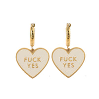A pair of heart-shaped earrings with the phrase 'FUCK YES' written across the front in a bold, white enamel against a gold hoop.