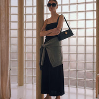 Woman in a black dress and sunglasses holding a black handbag in a room with large windows.