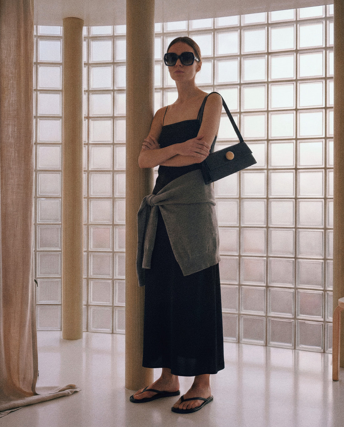 Woman in a black dress and sunglasses holding a black handbag in a room with large windows.