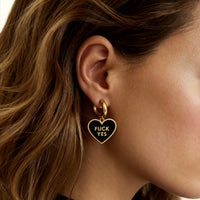 Heart-shaped earring with 'FUCK YES' text worn by a person with wavy hair.